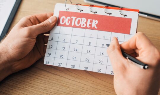 Cropped View Of Man Holding Pen And Calendar With October Month Near Laptop On Table